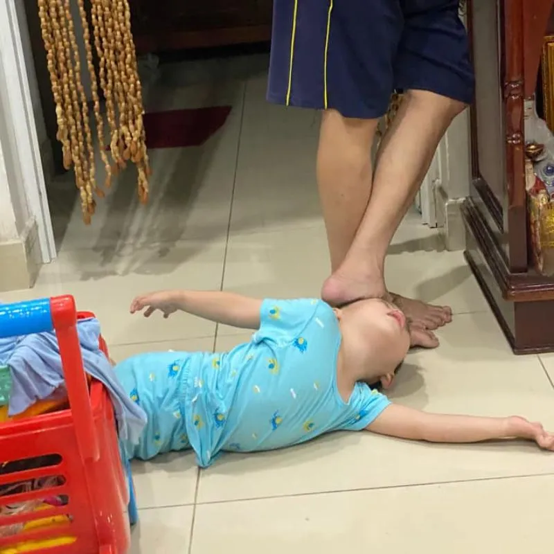 Em bé nằm ngửa trên sàn nhà, bị một bàn chân người lớn đặt lên mặt