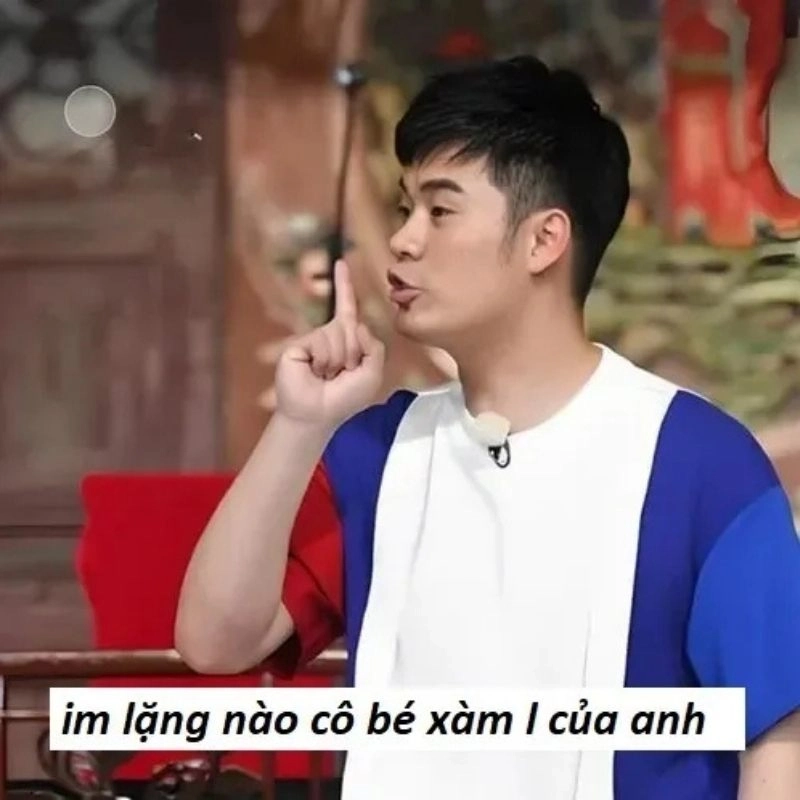Nam thanh niên mặc áo phối màu giơ ngón trỏ lên cao dặn im lặng trên truyền hình